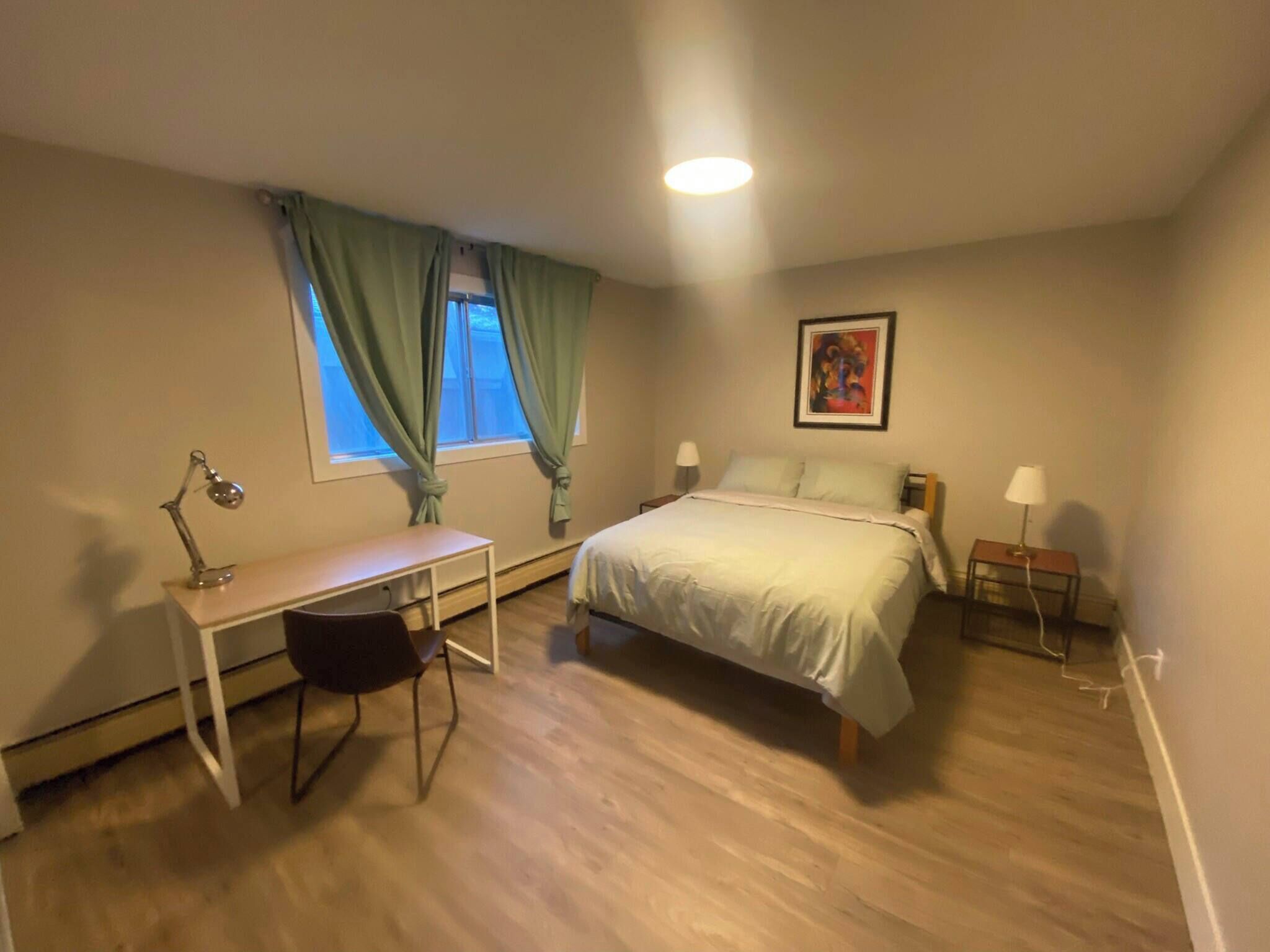 A bedroom with a bed, desk, chair, two bedside lamps, a window with curtains, and a painting on the wall.