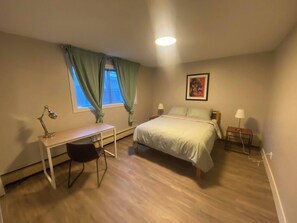 Un dormitorio con cama, escritorio, silla, dos lámparas de noche, ventana con cortinas y un cuadro en la pared.
