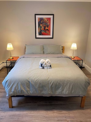 A neatly made bed with a striped bedspread, two pillows, and a folded towel set on the bed. There are two bedside lamps on wooden nightstands, and a framed picture hangs on the wall above the bed. - Brand New Modern Flat 2br1ba Whyte And U (Edmonton)