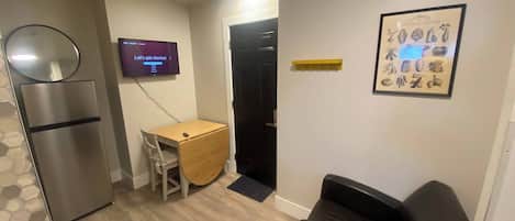 A compact living space with a kitchenette, a small desk, a black chair, and a framed picture on the wall.
