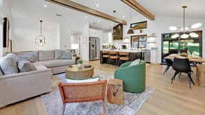 A spacious living room with a sofa, a coffee table, and a green armchair. A kitchen with a dining area is visible in the background. - Eugene Evergreen Escape by Les Chalets Stays is a fully renovated home perfect for your crew! Game room, play house, sauna, outdoor entertaining space and huge back yard. (Eugene)