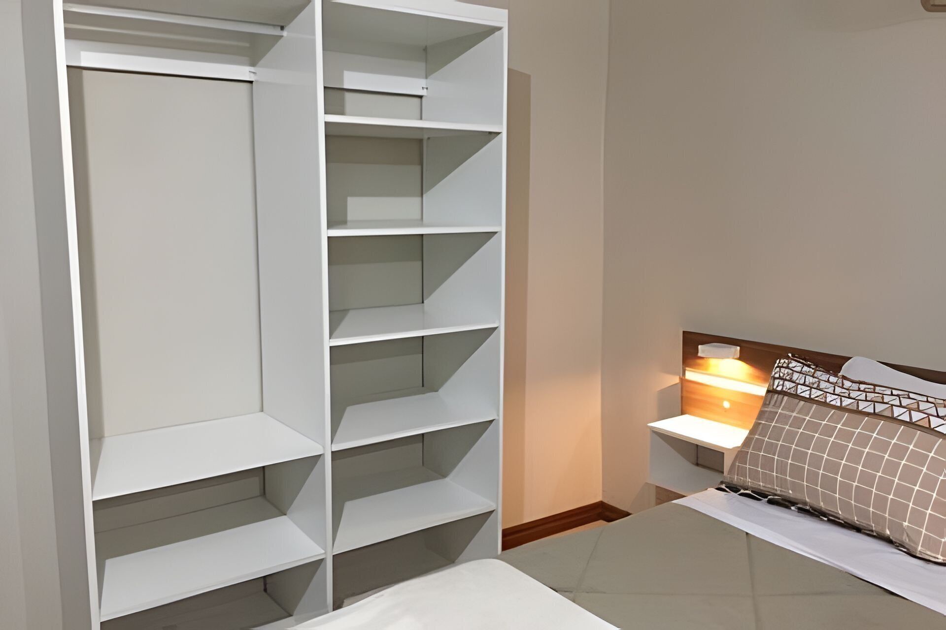 A bedroom with a white bed, a nightstand with a lamp, and a white shelving unit against the wall.