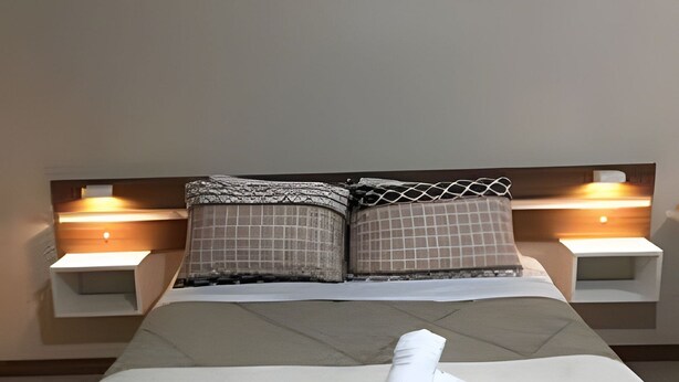 Una cama bien hecha con edredón gris, dos almohadas y una toalla blanca enrollada. Hay dos mesitas de noche con lámparas y un aire acondicionado de pared sobre la cama.