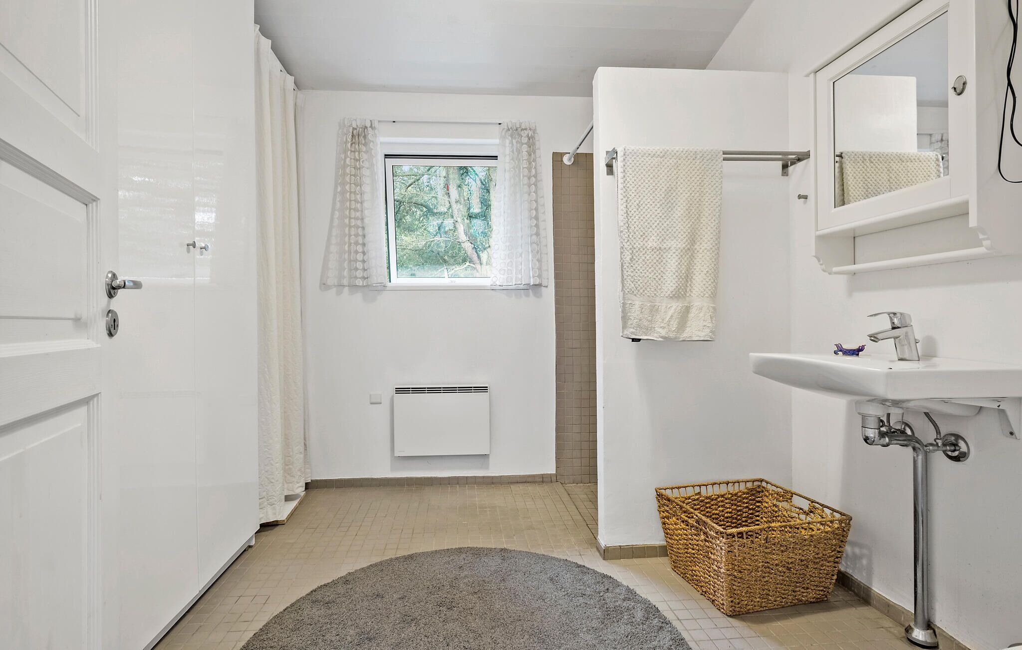 Une salle de bain avec une porte blanche, une fenêtre avec des rideaux, un porte-serviettes, un lavabo avec un miroir et un panier posé sur le sol.