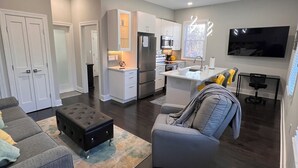 A modern living room with a grey sofa, a black ottoman, and a grey armchair. There is a kitchen area with white cabinets and a large flat-screen TV mounted on the wall. - Peaceful and modern retreat (Livingston)