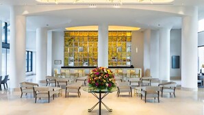 Un hall d’entrée spacieux, avec une table ronde centrale agrémentée de fleurs, flanqué de coins salon et, au fond, d’une grande vitrine illuminée.