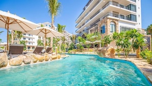Un espace piscine agrémenté de chaises longues et de parasols, entouré de palmiers et bordé d’un bâtiment doté de balcons.