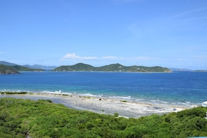 A coastal landscape with a beach, clear blue sea, and green hills in the background.