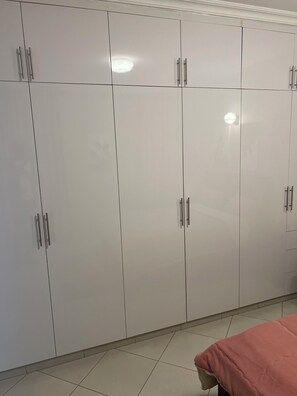A spacious room features a large wardrobe with multiple doors and handles, a bed with a cozy blanket, and a light-colored tile floor.