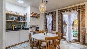 Een keuken met witte kasten, een ronde eettafel en houten stoelen. Er is een hanglamp en een bakstenen muur.