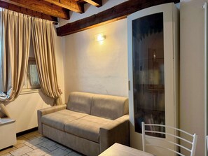 A room with a beige sofa, a white chair, a window with beige curtains, and a door with a glass panel. - San Bernardino 8 - Immobili e Soluzioni Rent (Bergamo)
