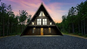 A modern, triangular-shaped house with a central entrance, large windows, and a gravel driveway, surrounded by a forest. - Hot Tub, X Box, Fire Pit, Close 2 Town/Lake Nearby, Pets, TH (Broken Bow)