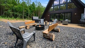 An outdoor seating area with benches, chairs, and a fire pit, surrounded by a forested landscape and a large building with a distinctive triangular roof. - Hot Tub, X Box, Fire Pit, Close 2 Town/Lake Nearby, Pets, TH (Broken Bow)