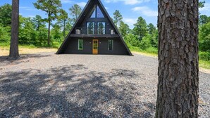 A modern, triangular wooden cabin with a balcony, surrounded by trees and gravel.