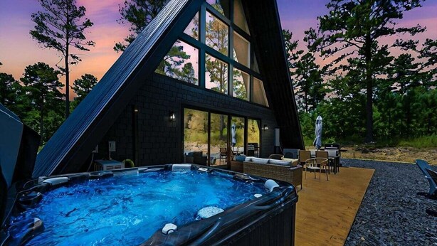A hot tub in front of a modern house with large windows, surrounded by outdoor furniture and a wooden deck.