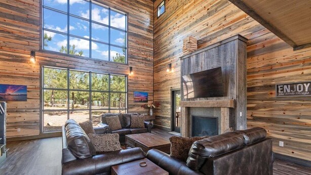 A rustic cabin interior with a leather sofa, wooden walls, large windows, and a fireplace. - Mountain Views, Hot Tub, Arcade, Pets, More Than A View (Broken Bow)
