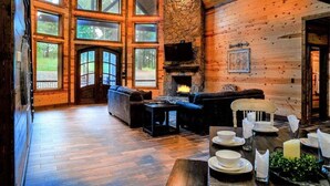 A cozy cabin interior with a stone fireplace, wooden walls, and large windows offering a view of the outdoors. - Arcade, Play Set, Hot Tub, Pets, Makin Plans (Broken Bow)