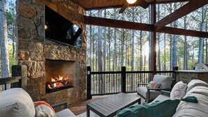 A stone fireplace with a TV mounted above it, surrounded by outdoor seating with cushions and a wooden table. - Fenced Yard, Hot Tub, Fire pit, Swings, Pets, Surfing Sunsets (Broken Bow)