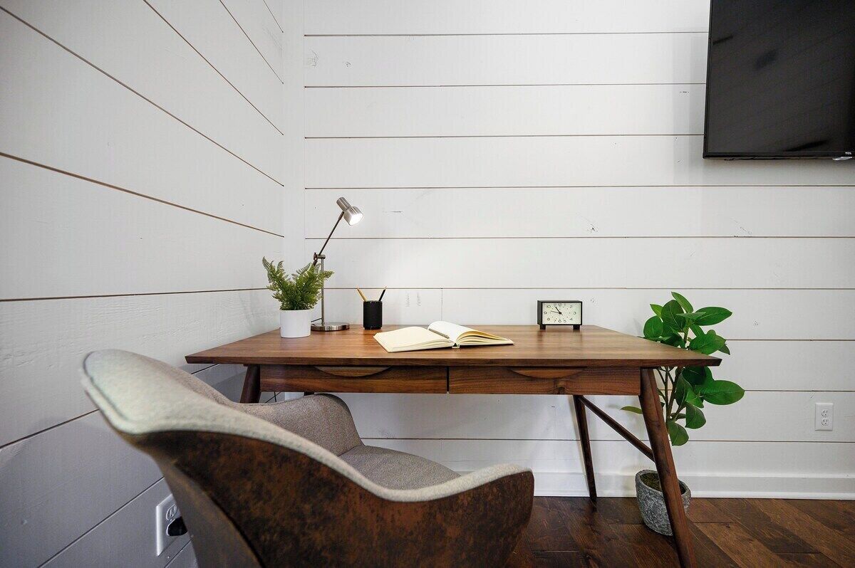A wooden desk with a lamp, a potted plant, and a book. A chair is positioned in front of the desk.