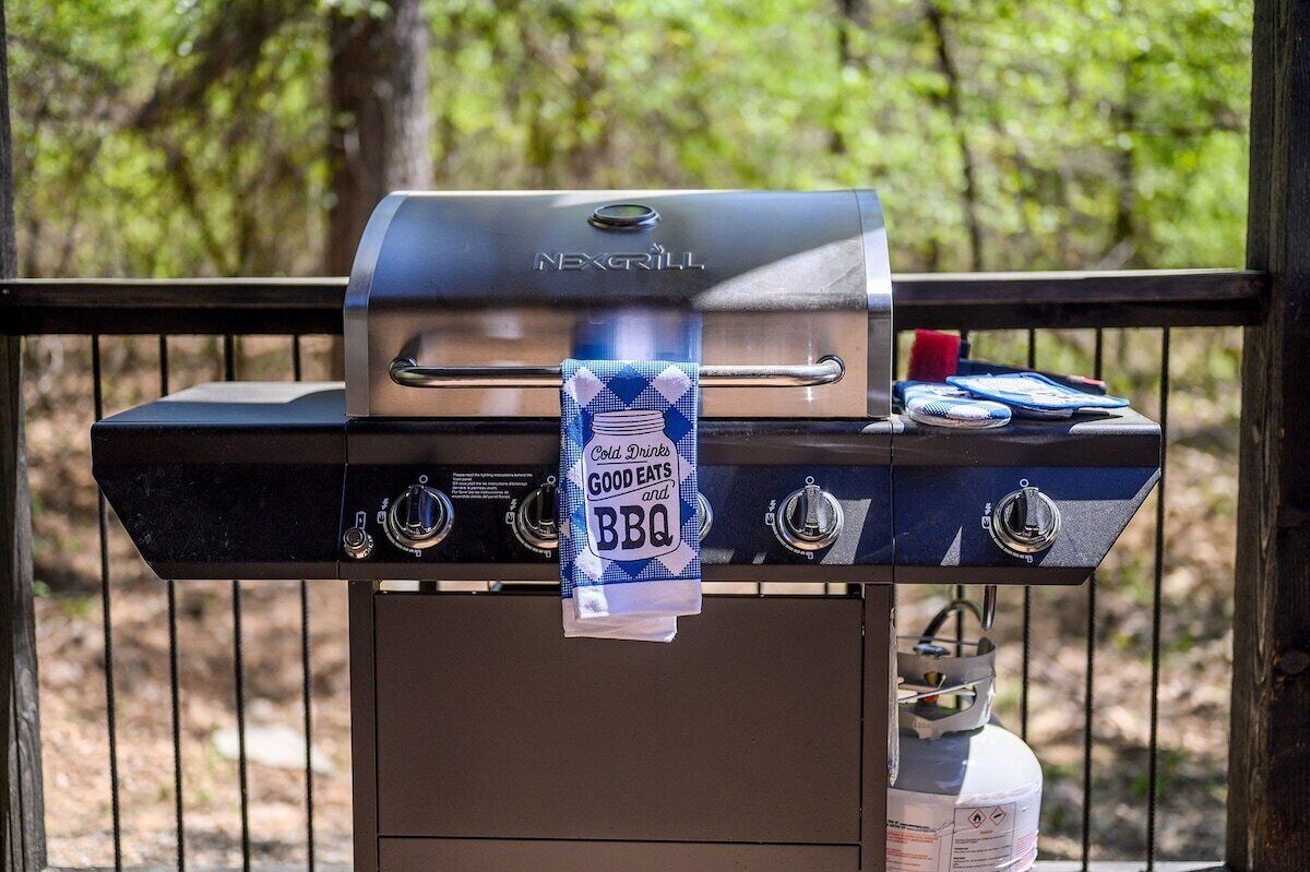 Una barbacoa NEXGRILL amb una tovallola que diu 'Bebidas fresques, menjar bo i barbacoa', i un dipòsit de propà a un costat.