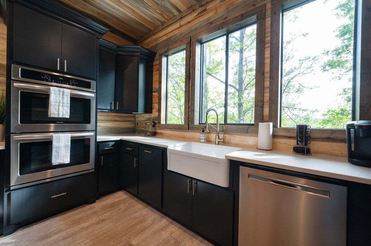 A modern kitchen with dark cabinetry, stainless steel appliances, and a large window offering a view of trees.