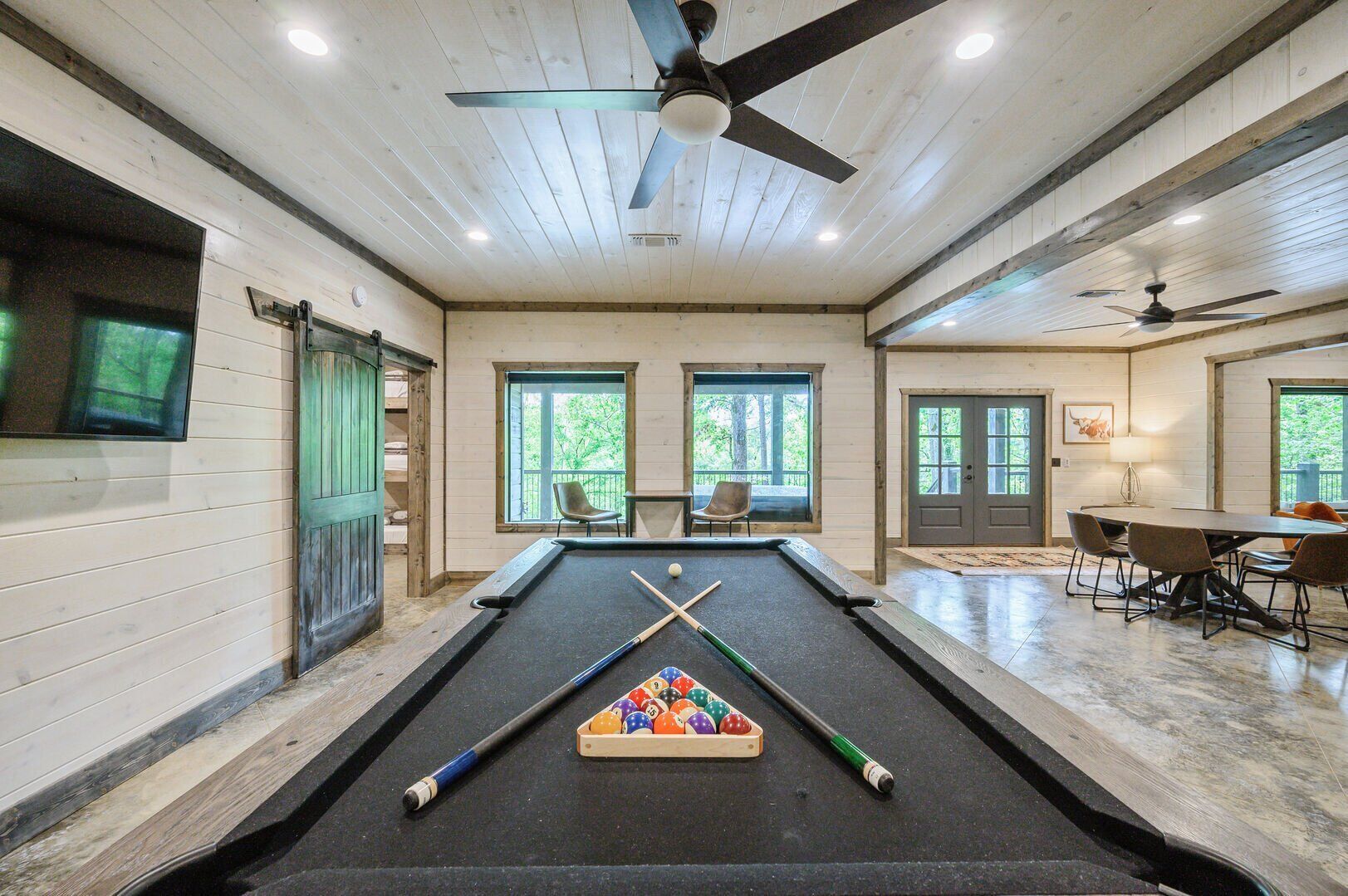 A pool table in a room with wooden walls and ceiling, two windows, and a ceiling fan.