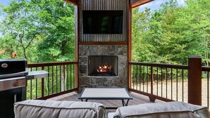 Une terrasse dotée d’une cheminée en pierre, d’un barbecue extérieur et d’un espace salon offrant une vue sur une forêt.