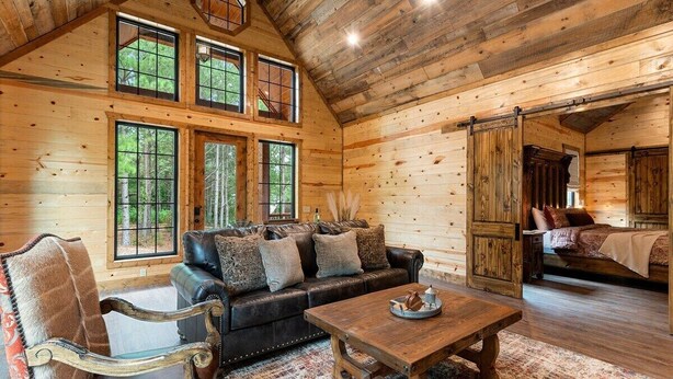 L’intérieur d’une cabane en rondins est aménagé avec un canapé en cuir, une table basse en bois et de grandes fenêtres qui offrent une vue sur la forêt.