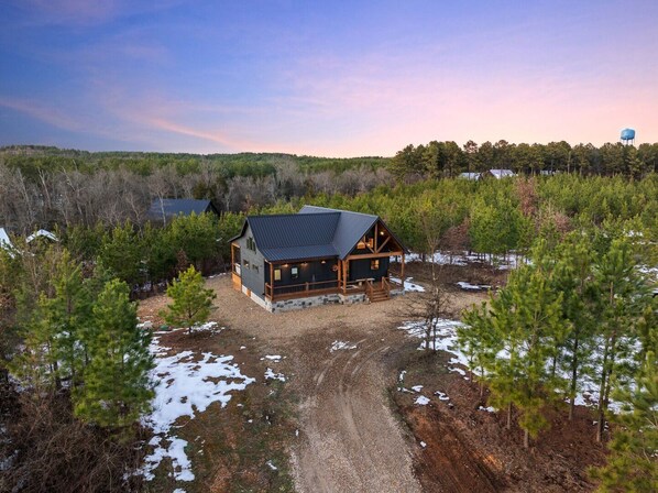 A cabin with a stone foundation, a metal roof, and a wooden deck. - Hot Tub, Soaking Tub, Romantic, Close 2 Town, Pets, Vacana Pine & Bow (Broken Bow)