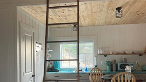 A kitchen with a wooden ladder, white cabinets, a window, and a dining area with wooden chairs. - COZY CABIN. One room cabin with loft. Great sunsets & river view before Spring! (Great Cacapon)