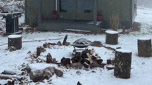 A wooden cabin with a covered porch, a fire pit, and a snow-covered landscape. - COZY CABIN. One room cabin with loft. Great sunsets & river view before Spring! (Great Cacapon)