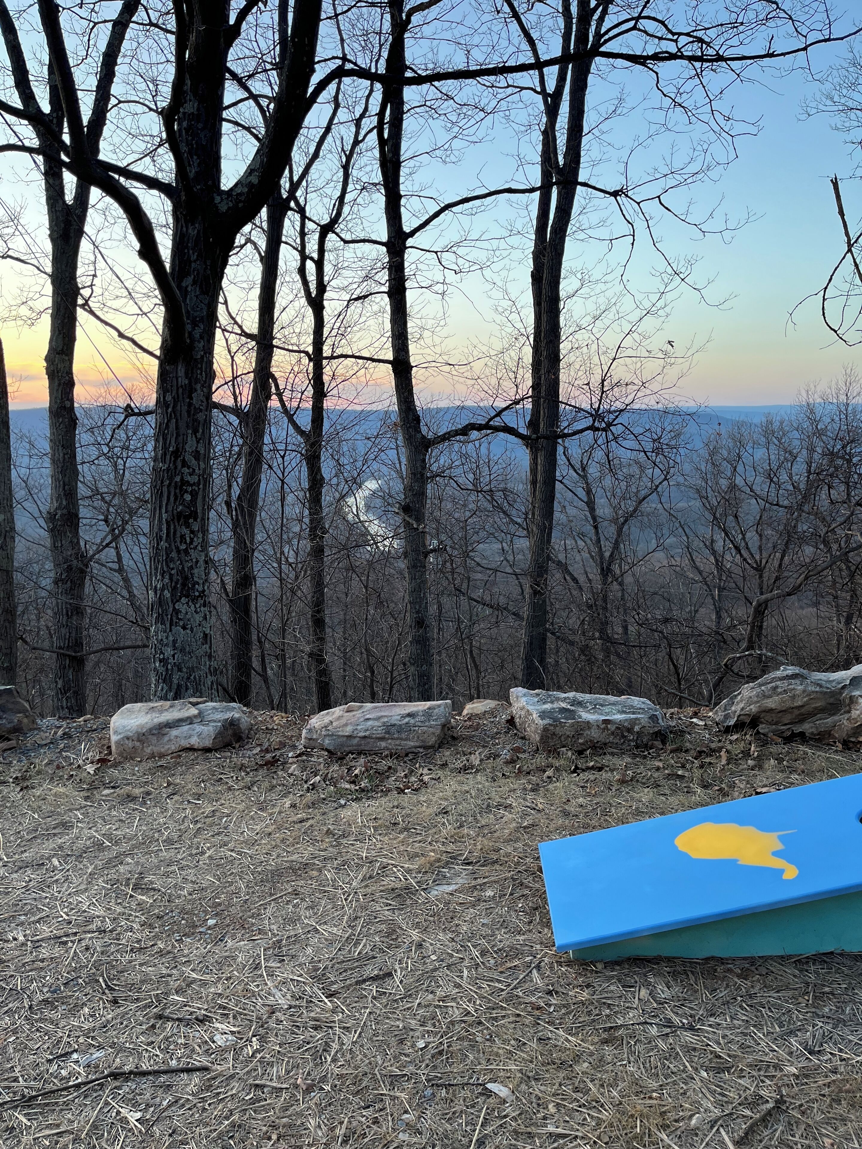 Vista de una zona boscosa con cielo despejado y un atardecer. Se observan rocas grandes y una esterilla azul con un diseño amarillo en el suelo.