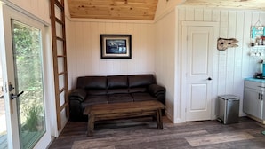 A room with a wooden ceiling, a brown leather sofa, a wooden bench, a white door, and a small kitchen area with a trash bin. - COZY CABIN. One room cabin with loft. Great sunsets & river view before Spring! (Great Cacapon)