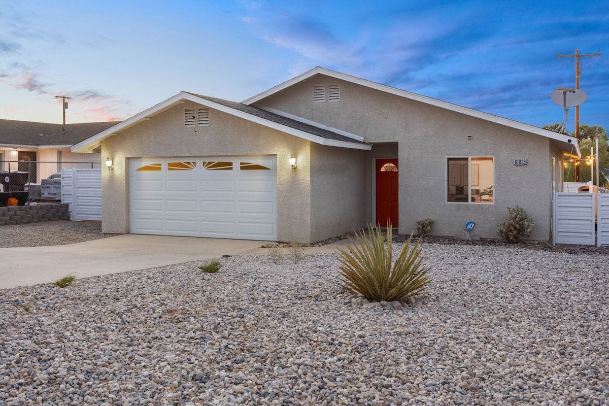 A single-story house with a garage, a red door, and a small garden with desert plants.