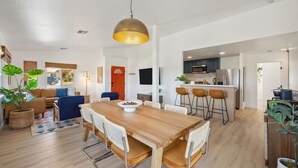 A modern kitchen with a central island, a dining area with a wooden table and chairs, and a living room with a sofa and a TV. - Tangerine House: Close to Downtown JT, Hot Tub & Game Room (Joshua Tree)