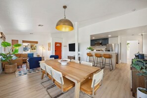 A modern kitchen with a central island, a dining area with a wooden table and chairs, and a living room with a sofa and a TV. - Tangerine House: Close to Downtown JT, Hot Tub & Game Room (Joshua Tree)