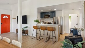 A modern kitchen with bar stools, a red door, and a dining area with a table and chairs. - Tangerine House: Close to Downtown JT, Hot Tub & Game Room (Joshua Tree)