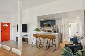 A modern kitchen with bar stools, a red door, and a dining area with a table and chairs. - Tangerine House: Close to Downtown JT, Hot Tub & Game Room (Joshua Tree)