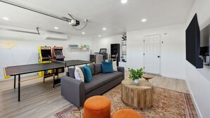 A modern living room with a ping pong table, sofa, ottomans, and a flat-screen TV. - Tangerine House: Close to Downtown JT, Hot Tub & Game Room (Joshua Tree)