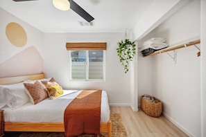 A bedroom with a bed, a window with blinds, a ceiling fan, a hanging plant, and a shoe rack.