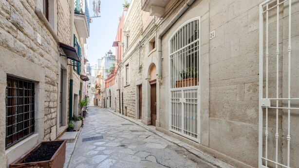 A narrow stone-paved street flanked by historic buildings with barred windows and doors, and a number '24' visible on one building. - GuestHost - Albachiara Home in Andria (Andria)
