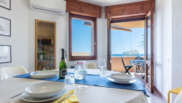 Ein Esstisch ist gedeckt mit Tellern, Gläsern und Besteck, einer Flasche und einer Kanne, mit Blick auf das Meer durch offene Türen.