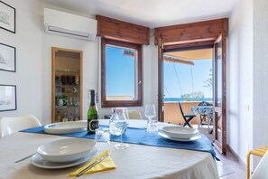 A dining table set with plates, glasses, and cutlery, a bottle, and a pitcher, with a view of the sea through open doors.