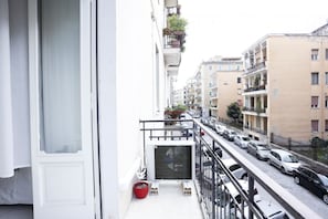 Una calle estrecha con coches aparcados, un balcón con una maceta roja y edificios de varios pisos con balcones.