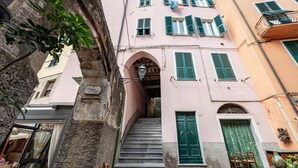 Eine schmale Straße mit einem rosa Gebäude, das eine Treppe, grüne Fensterläden und ein Geschäft im unteren Bereich zeigt.