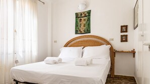 Ein ordentlich bezogenes Bett mit weißen Leinen und aufgerollten Handtüchern, ein Korbschlafzimmer, eine wandmontierte Lampe und eine kleine Ablage mit Bildern.
