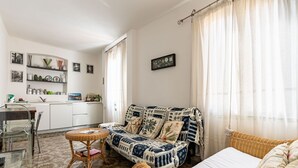 Ein gemütliches Wohnzimmer mit einer Couch, einem kleinen Tisch und einem Korbstuhl. Es gibt eine Küchenzeile mit Schränken und einer Arbeitsplatte.