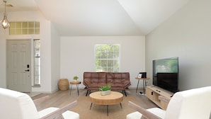 A modern living room with a brown leather sofa, white armchairs, a wooden coffee table, and a large flat-screen TV. - SeaWorld San Antonio! Quiet, 4-bedroom house. 12 guests. (San Antonio)