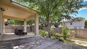 A covered patio with a wooden deck, a table and chairs, and a small garden area with a tree and shrubs. - SeaWorld San Antonio! Quiet, 4-bedroom house. 12 guests. (San Antonio)