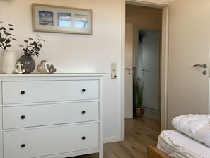 Un dormitorio con un aparador blanco, una cama con ropa de cama blanca, un cuadro en la pared y una planta en maceta en la esquina.